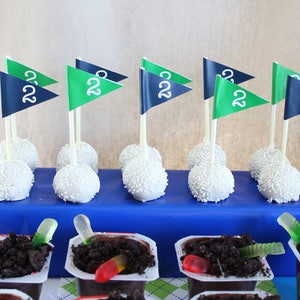 May include: A blue and green golf-themed cake pops with white frosting and sprinkles. The cake pops are arranged on a blue surface. There are also chocolate pudding cups with gummy worms on top.