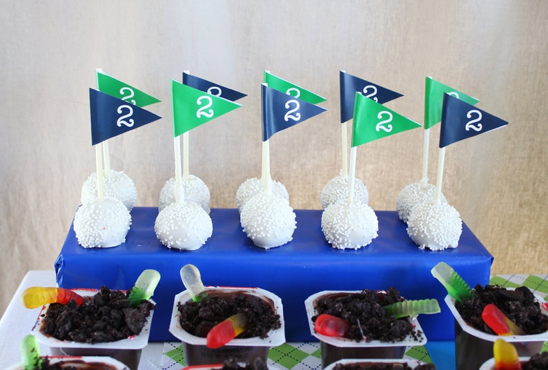 May include: A blue and green golf-themed cake pops with white frosting and sprinkles. The cake pops are arranged on a blue surface. There are also chocolate pudding cups with gummy worms on top.