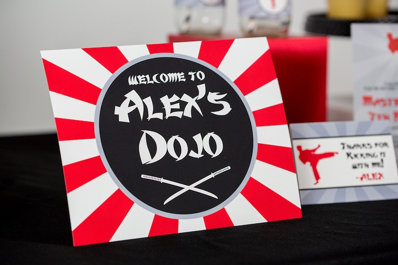 May include: A red and white striped sign with a black circle in the center. The circle says "Welcome to Alex's Dojo" in white text with two crossed swords underneath.