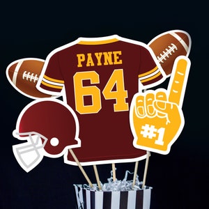 Puede incluir: Centro de mesa con temática de fútbol con una camiseta granate y dorada, un balón de fútbol, un casco y una mano de espuma. La camiseta tiene el nombre "PAYNE" y el número "64" en la espalda. La mano de espuma tiene el número "#1" en ella.