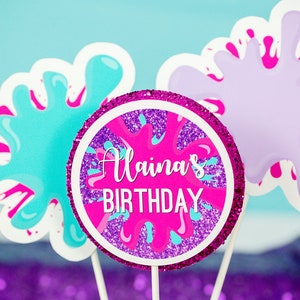 May include: Three pink, purple, and blue paint splatter cake toppers with a pink circle topper that says "Alaina's Birthday" in white text.