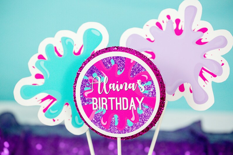 May include: Three pink, purple, and blue paint splatter cake toppers with a pink circle topper that says "Alaina's Birthday" in white text.