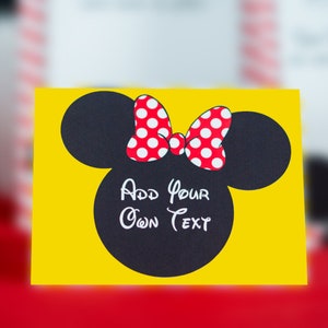 Peut inclure: Une carte jaune avec une silhouette noire de la t&ecirc;te de Minnie Mouse avec un n&oelig;ud rouge &agrave; pois blancs. Le texte "Add your own text" est &eacute;crit en blanc &agrave; l'int&eacute;rieur de la silhouette.