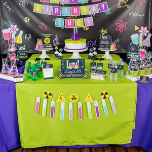 Pu&ograve; includere: Un allestimento per una festa di compleanno a tema scientifico con uno schema di colori verde e viola. Lo striscione recita "Happy 8th Birthday Lucy". Il tavolo &egrave; decorato con becher, provette e una torta. Il tema include elementi "Slime Station" e "Biohazard".