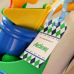May include: A green and blue golf bag with a yellow and orange golf club and a golf ball inside. A tag with a green, white, and blue diamond pattern is attached to the bag. The tag says "this bag belongs to: Anthony".
