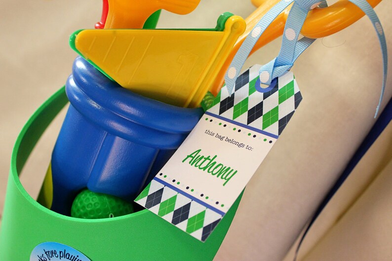 May include: A green and blue golf bag with a yellow and orange golf club and a golf ball inside. A tag with a green, white, and blue diamond pattern is attached to the bag. The tag says "this bag belongs to: Anthony".