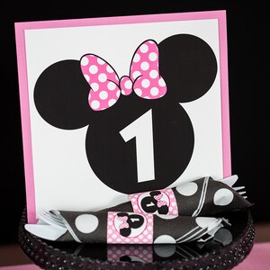 May include: A pink and black Minnie Mouse themed table number card with the number "1" in the center. The card is on a black cake stand with black and white polka dot napkins.