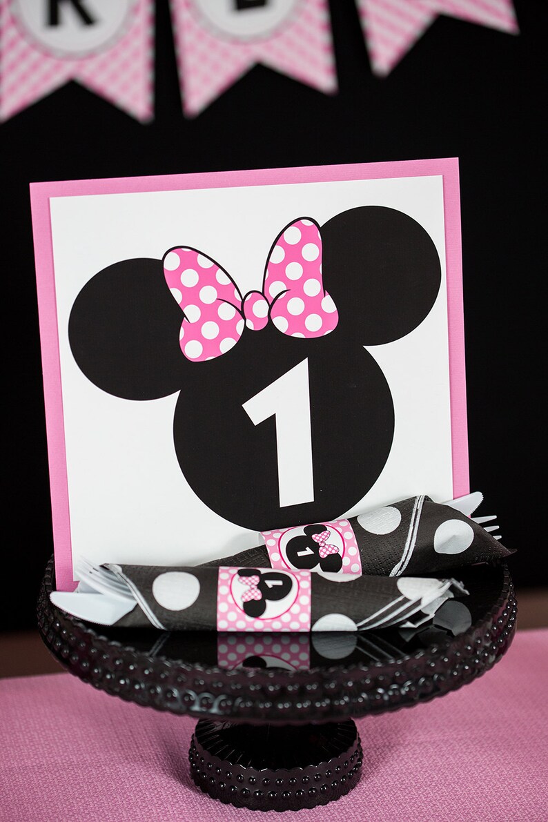 May include: A pink and black Minnie Mouse themed table number card with the number "1" in the center. The card is on a black cake stand with black and white polka dot napkins.