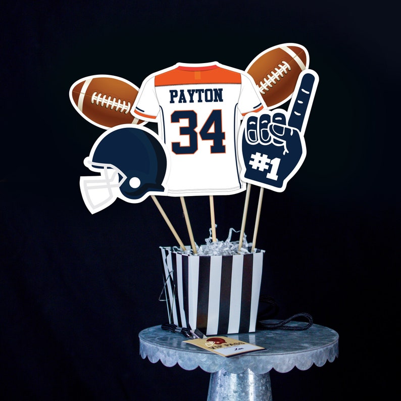 May include: A set of football-themed party decorations, including a football jersey with the name "PAYTON" and the number "34", a football helmet, a football, a foam finger with the number "#1", and a black and white striped container.