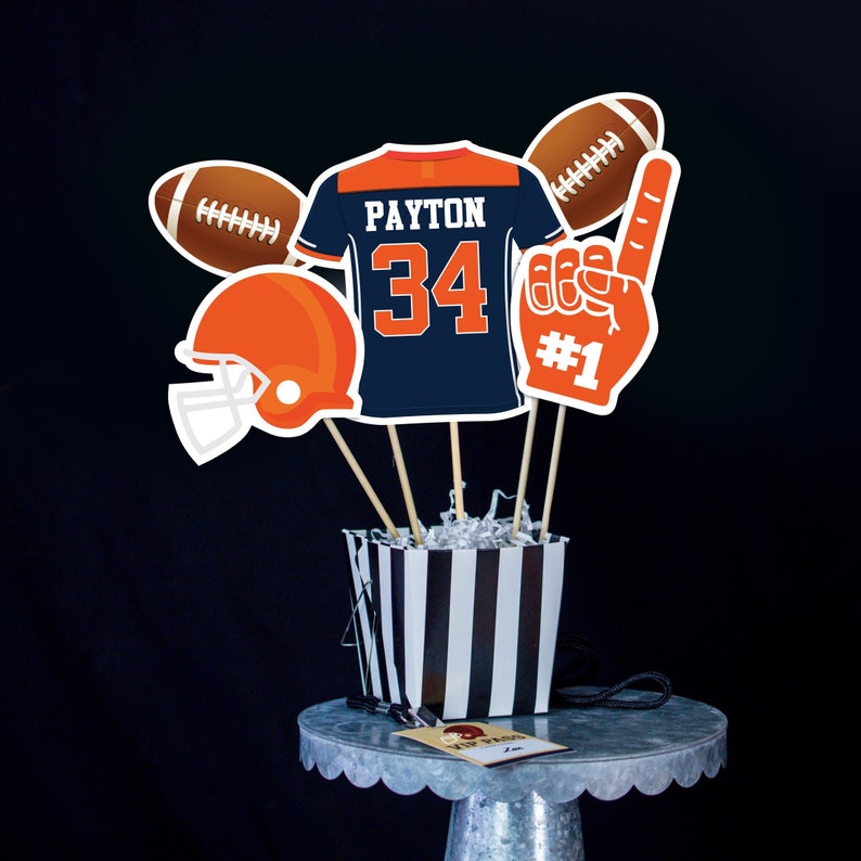 May include: A football-themed centerpiece with a blue and orange jersey with the name "PAYTON" and the number "34" on it. The centerpiece also includes a football, a helmet, a foam finger, and a black and white striped container.