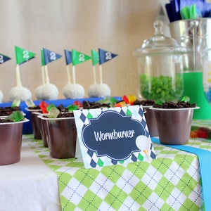 May include: A golf-themed dessert table with chocolate pudding cups topped with gummy worms and a sign that reads "Wormburner". The table is covered in a green and white checkered tablecloth.