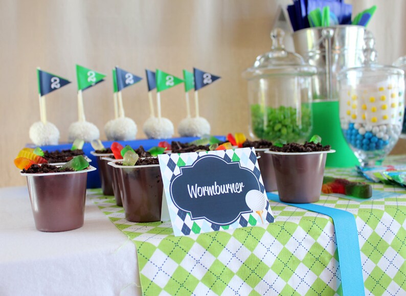 May include: A golf-themed dessert table with chocolate pudding cups topped with gummy worms and a sign that reads "Wormburner". The table is covered in a green and white checkered tablecloth.