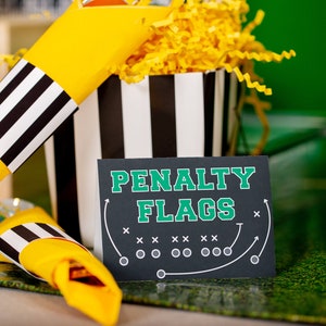 Puede incluir: Un cono de papel a rayas blanco y negro lleno de papel amarillo triturado. Un letrero negro con texto blanco dice "PENALTY FLAGS" con un gr&aacute;fico de campo de f&uacute;tbol verde.