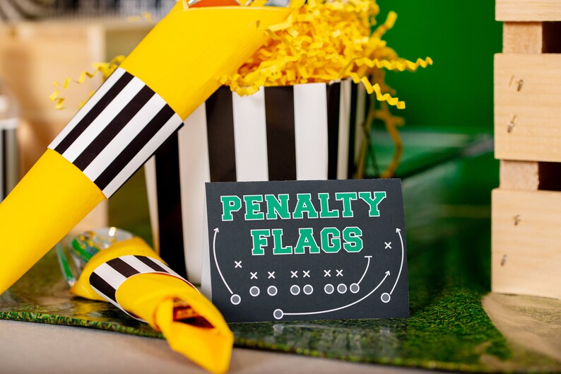 Puede incluir: Un cono de papel a rayas blanco y negro lleno de papel amarillo triturado. Un letrero negro con texto blanco dice "PENALTY FLAGS" con un gr&aacute;fico de campo de f&uacute;tbol verde.