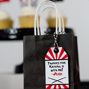 May include: Black and red party favor bag with a white handle and a red and white tag that says "Thanks for kicking it with me! -Alex".