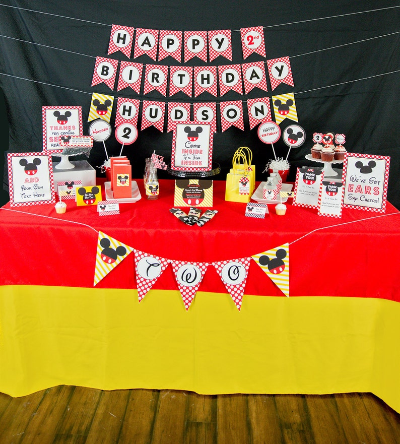 May include: A festive birthday party setup with a red and yellow tablecloth. Decorations include banners, signs, and party favors featuring a cartoon mouse character. The banner reads "Happy Birthday Hudson 2".