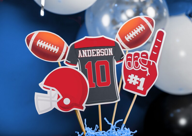 Puede incluir: Un centro de mesa con tem&aacute;tica de f&uacute;tbol con una camiseta roja y negra con el nombre "Anderson" y el n&uacute;mero "10" en ella, un casco de f&uacute;tbol rojo y blanco, un bal&oacute;n de f&uacute;tbol rojo y blanco, un dedo de espuma rojo y blanco con el n&uacute;mero "#1" en &eacute;l, y un bal&oacute;n de f&uacute;tbol rojo y blanco.