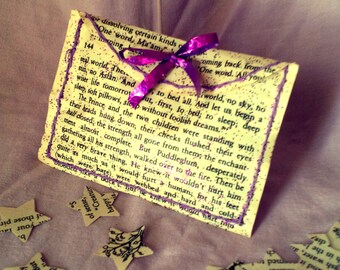 May include: A folded piece of paper with a purple ribbon tied around it. The paper has text printed on it, including the words "One word, Ma'am" and "The teal world." The paper is sitting on a purple and white fabric background.