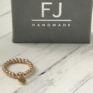 May include: A gold ring with a small heart charm hanging from it. The ring is made of small gold beads. The ring is on a white wooden surface. The ring is in a box with the text "FJ HANDMADE" on it.