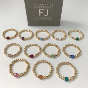 May include: Twelve gold bead rings with a single crystal bead in various colours, including red, purple, blue, green, and orange. The rings are arranged in three rows of four. The rings are displayed on a white surface. The image also includes a box with the text "FUNKSION JEWELLERY FJ HANDMADE".