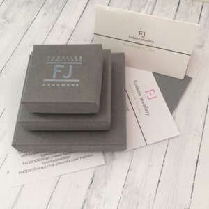May include: A gray box with the text "FUNKSION JEWELLERY FJ HANDMADE" on the top. The box is sitting on top of two other gray boxes. A white card with the text "FJ funksion jewellery handmade for you" is sitting on top of a gray envelope.
