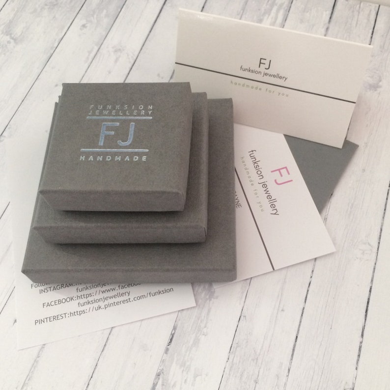 May include: A gray box with the text "FUNKSION JEWELLERY FJ HANDMADE" on the top. The box is sitting on top of two other gray boxes. A white card with the text "FJ funksion jewellery handmade for you" is sitting on top of a gray envelope.