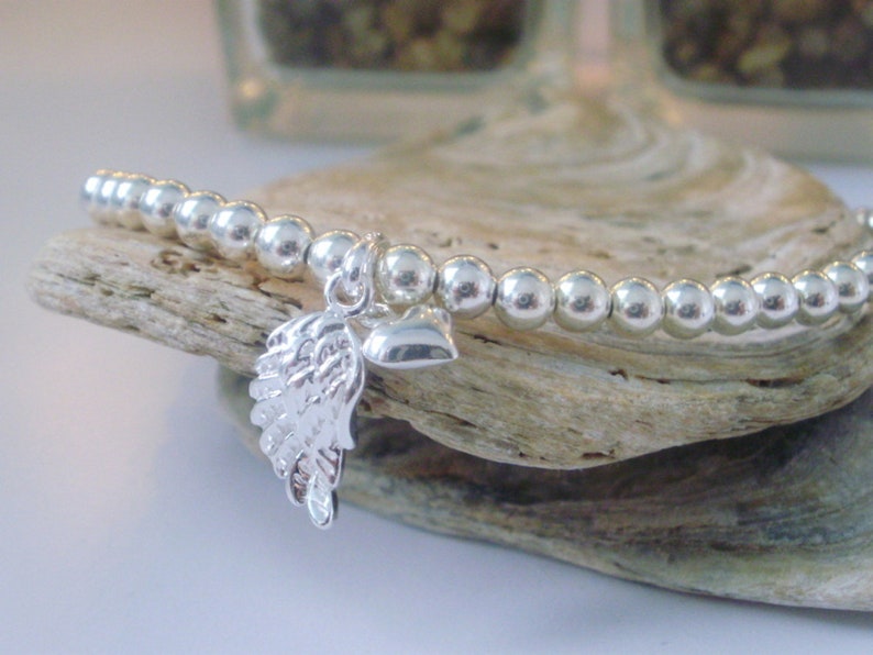 May include: A silver bracelet with a small silver heart and angel wing charm. The bracelet is made of small silver beads.