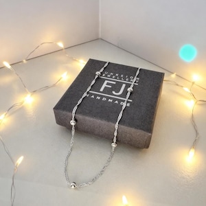 May include: A silver necklace featuring a twisted rope chain and spherical beads. The necklace is presented on a dark grey box with the text "UNKSION JEWELLERY" and "FJ HANDMADE". Fairy lights illuminate the background.