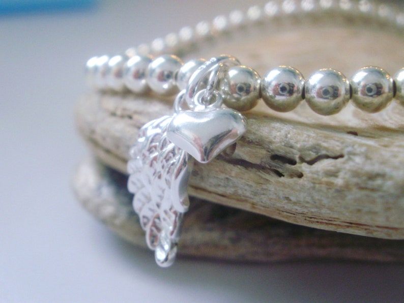 May include: A silver bracelet with a charm of a heart and a wing. The bracelet is made of small silver beads.