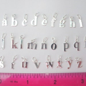 May include: A set of 26 silver letter charms, each charm is a different letter of the alphabet. The charms are arranged in alphabetical order on a white surface with a pink ruler for scale.