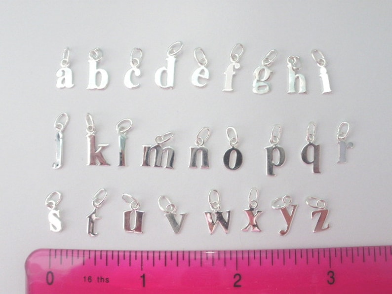 May include: A set of 26 silver letter charms, each charm is a different letter of the alphabet. The charms are arranged in alphabetical order on a white surface with a pink ruler for scale.