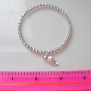 May include: A silver beaded bracelet with a small silver heart and angel wing charm. The bracelet is measured against a pink ruler.