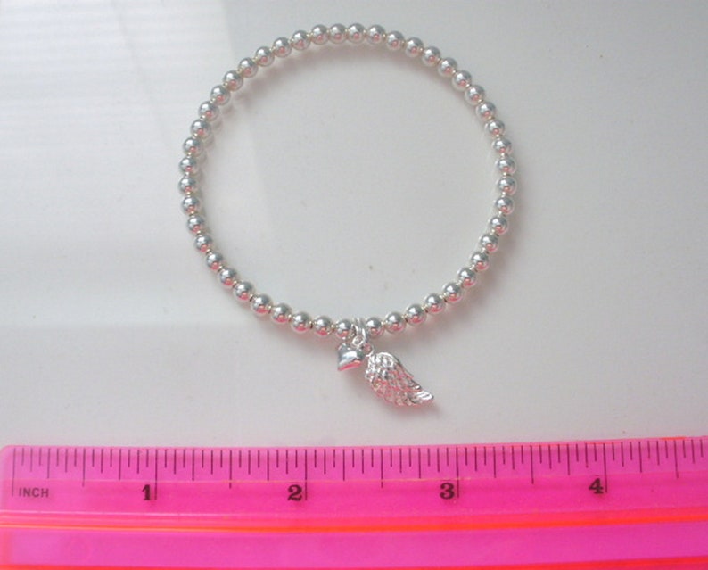 May include: A silver beaded bracelet with a small silver heart and angel wing charm. The bracelet is measured against a pink ruler.