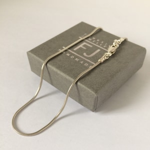 May include: A silver snake chain necklace with a lobster clasp. The necklace is in a grey box with the text "FJ Handmade Jewellery" printed on it.
