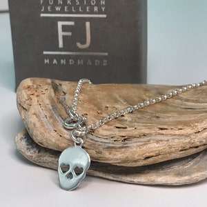 May include: Silver necklace featuring a skull charm. The skull has two heart-shaped cutouts for the eyes. The necklace is displayed on a wooden surface. The text "FJ Handmade" is visible on a box in the background.