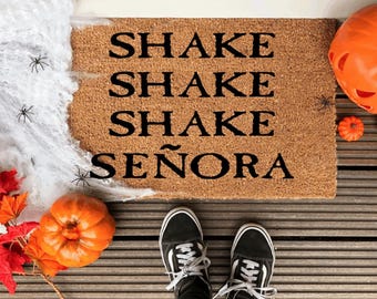 Puede incluir: Una alfombra de coco marrón con el texto "Shake Shake Shake Señora" en letras negras. La alfombra está sobre una superficie de madera con una telaraña y calabazas.