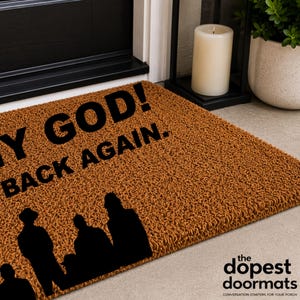 Puede incluir: Un felpudo rectangular de fibra de coco marr&oacute;n con el texto negro "OH MY GOD! THEY'RE BACK AGAIN." y las siluetas negras de cuatro figuras en fila. La alfombra tiene una superficie texturizada y fibrosa sobre un suelo claro.