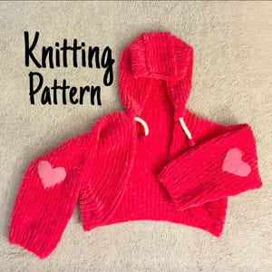 Könnte beinhalten: Ein leuchtend pinkfarbener Kapuzen-Bolero-Strickpullover mit einem hellrosa Herz auf jedem Ärmel. Die Worte "Knitting Pattern" sind in schwarzer Schrift geschrieben. Der Pullover hat weiße Kordelzüge.