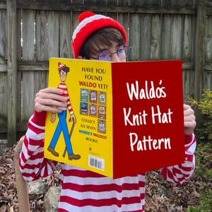 May include: A person wearing a red and white striped shirt and a red and white knitted hat is holding open a book titled "Waldo's Knit Hat Pattern". The book cover is red with white text. The book's back cover is yellow with a picture of Waldo and the text "Have you found Waldo yet?"
