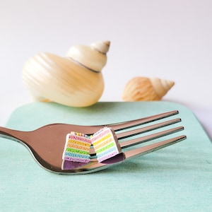 Könnte beinhalten: Ein Miniatur-Regenbogen-Kuchenstück auf einer silbernen Gabel. Der Kuchen ist mit Streuseln verziert und hat Schichten aus rosa, gelbem, grünem und blauem Frosting.