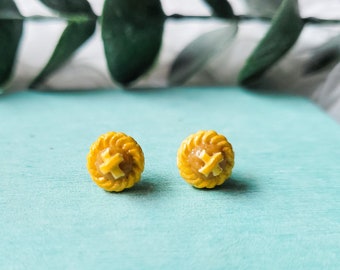 Pode incluir: Um par de brincos de torta em miniatura amarelos. Os brincos são em forma de crosta de torta com um recheio marrom.