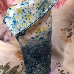 May include: A handmade ceramic vase with a unique, asymmetrical shape. The vase features a blend of colors, including blue, white, and yellow, with a textured, speckled pattern. The lower portion transitions to a darker blue hue, creating a gradient effect.