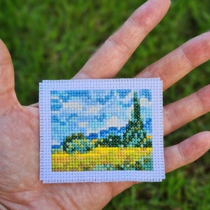 Può includere: Un piccolo ricamo a punto croce che rappresenta una scena di paesaggio con un cielo blu, nuvole bianche, alberi verdi e un campo giallo.