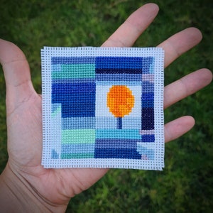 May include: A small, square cross-stitch piece with a geometric design featuring a blue, green, and orange color scheme. The center of the design is a bright orange circle with a small brown tree inside.