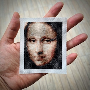 Puede incluir: Un bordado de punto de cruz enmarcado en blanco de un rostro. El rostro está realizado en hilo marrón, negro y blanco. Los ojos de la persona están cerrados y su boca está ligeramente abierta.