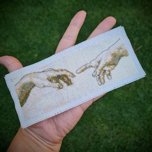 Puede incluir: Una bordado de punto de cruz blanco que representa dos manos que se alcanzan, inspirado en La Creación de Adán de Miguel Ángel. Las manos están detalladas con tonos de marrón, verde y amarillo.