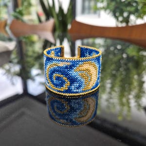 May include: A wide cuff bracelet crafted from tiny glass beads in shades of blue, white, and yellow. The beadwork features a swirling pattern of a crescent moon and stars, bordered by a thin line of metallic gold beads along the top and bottom edges.