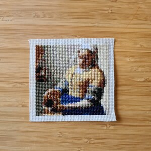 May include: A cross-stitch embroidery of a woman in a blue and white dress holding a brown jug. The woman is wearing a white cap and has brown hair. The background is a light brown.