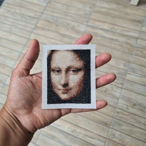May include: A white framed cross-stitch of a portrait of the Mona Lisa. The cross-stitch is done in shades of brown and black.