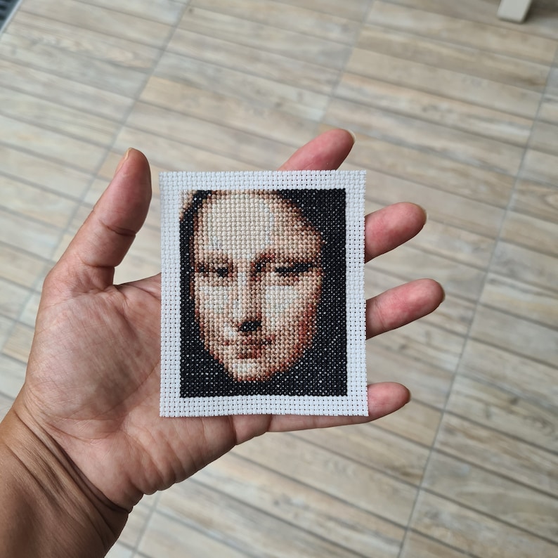 May include: A white framed cross-stitch of a portrait of the Mona Lisa. The cross-stitch is done in shades of brown and black.